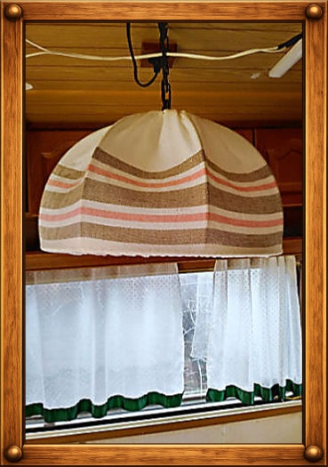 Hängelampe mit gestreiftem Stoffschirm über einem Fenster mit Gardine in einem Wohnwagen.