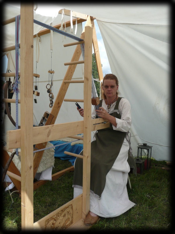 Handwerkerin in historischer Gewandung arbeitet an einem großen Holzwebstuhl in einem mittelalterlichen Lagerzelt.