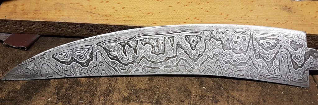 Damastklinge ohne Griff mit markanter Musterung auf Werkbank.